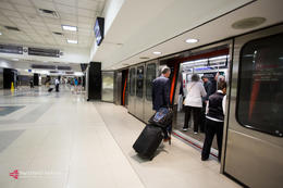 You can see the boarding platform of an airport shuttle. The floor is tiled with large light-coloured tiles. A passenger in a suit is preparing to cross the threshold of the shuttle, on the right, dragging behind him a wheeled suitcase and a backpack. Other passengers have already boarded the shuttle, whose doors are open, as are the station’s platform doors. Other passengers can be seen waiting or moving in the background. Overhead signage can be seen at the top as well as a video screen above the entrance